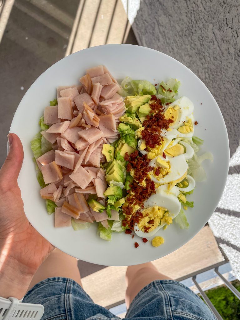 Hand holding a plate of cobb salad with chopped turkey, avocado, hard boiled egg, and bacon bits.