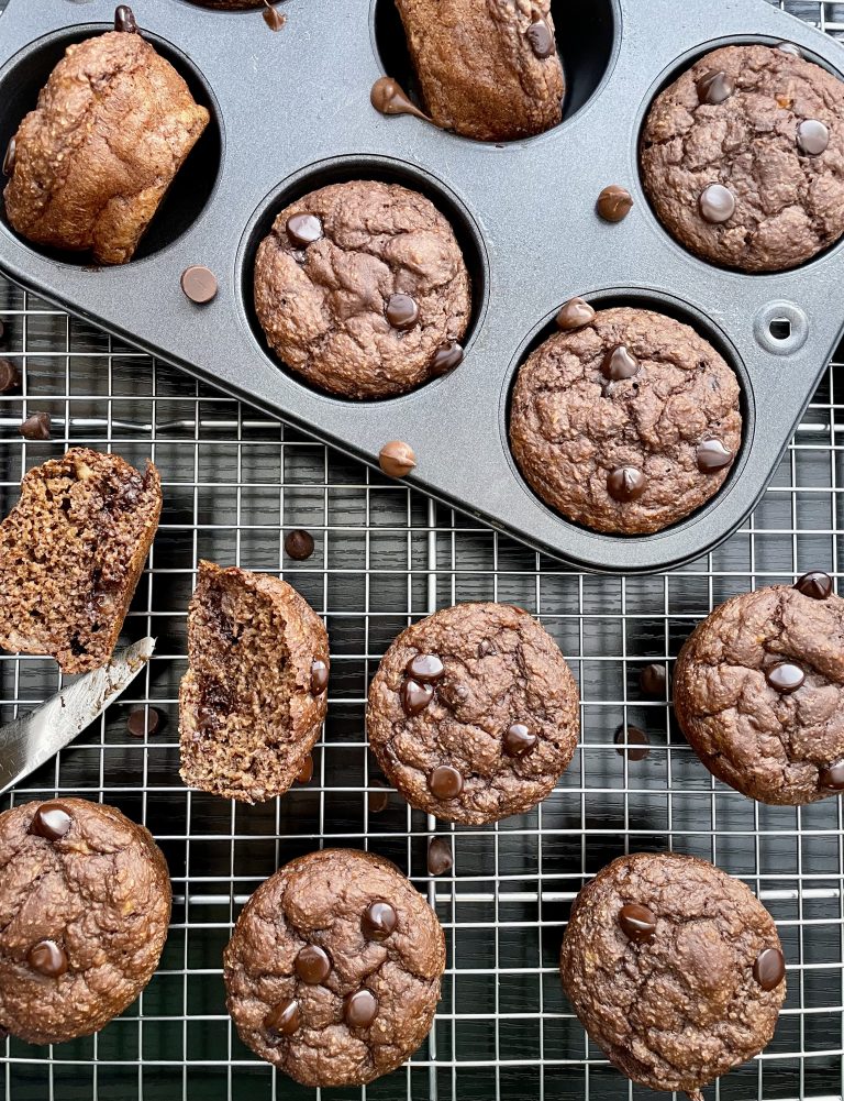Chocolate banana oat muffins with chocolate chips in a muffin tin, on a cooling rack, with one cut open with a butter knife.