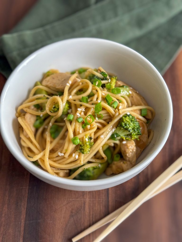 Bowl of chicken and broccoli lo mein with green onion and sesame seeds, chopsticks and green kitchen towel in the background.