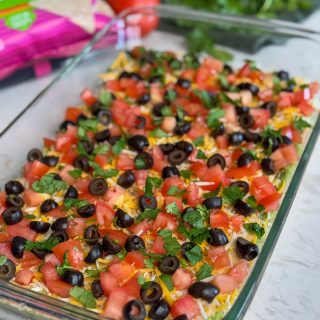 Glass dish of easy 7 layer dip with tomatoes, olives, cheese and cilantro.