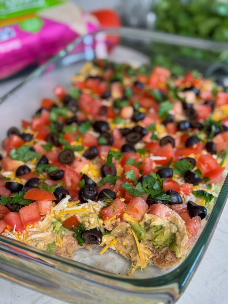 Glass dish of easy 7 layer dip with tomatoes, olives, cheese and cilantro.
