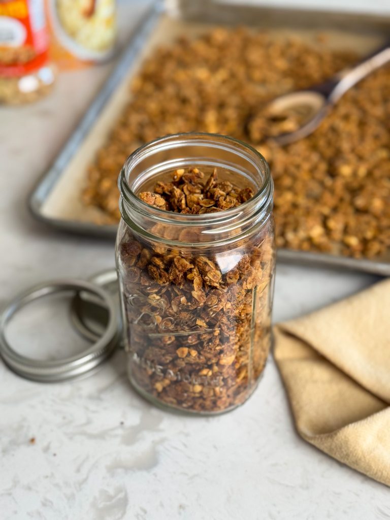 Healthy homemade peanut butter granola in a large mason jar with sheet pan of granola in the background.