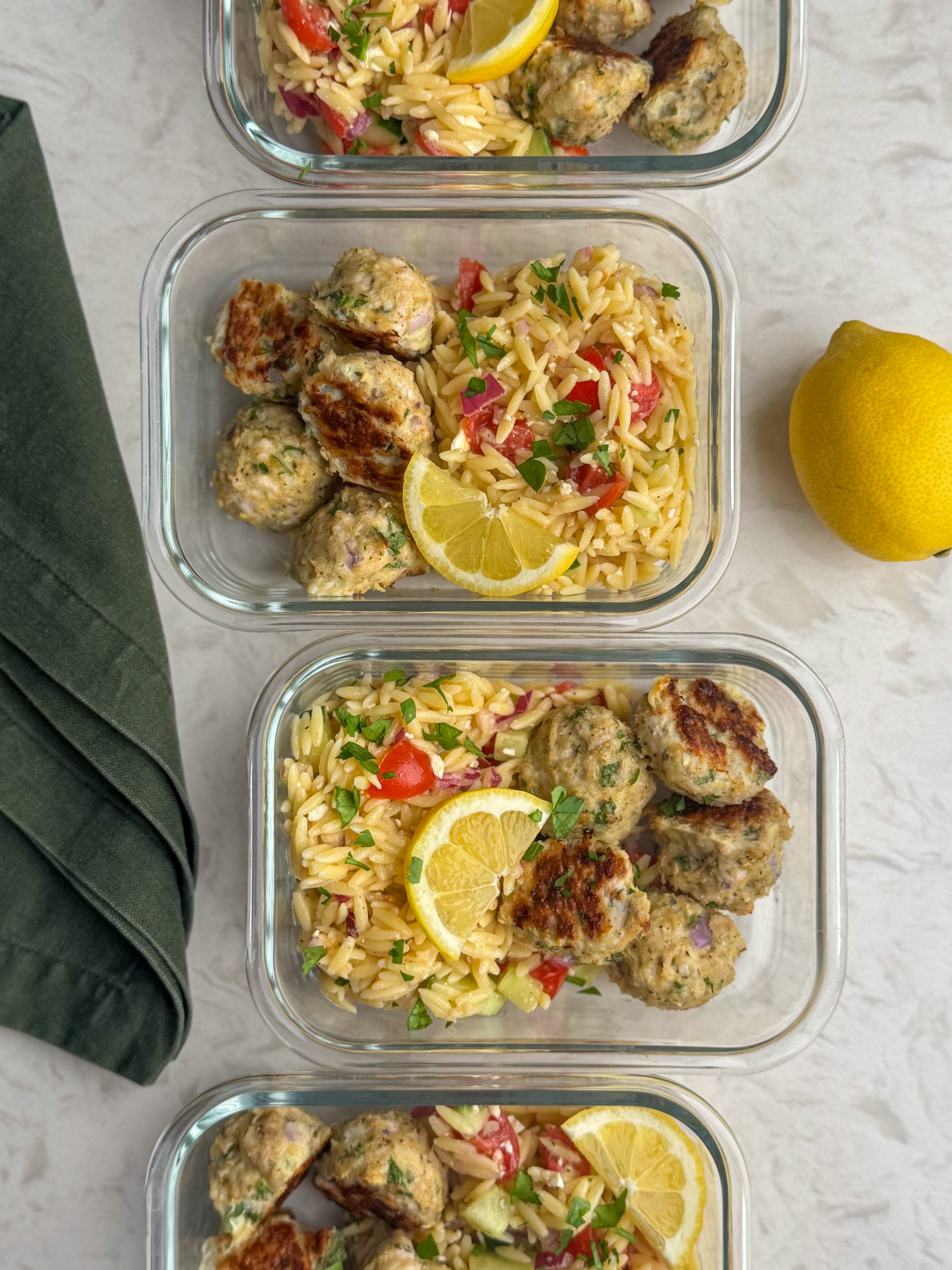 Greek turkey meatballs with orzo salad in glass containers with fresh lemon slices and green towel.