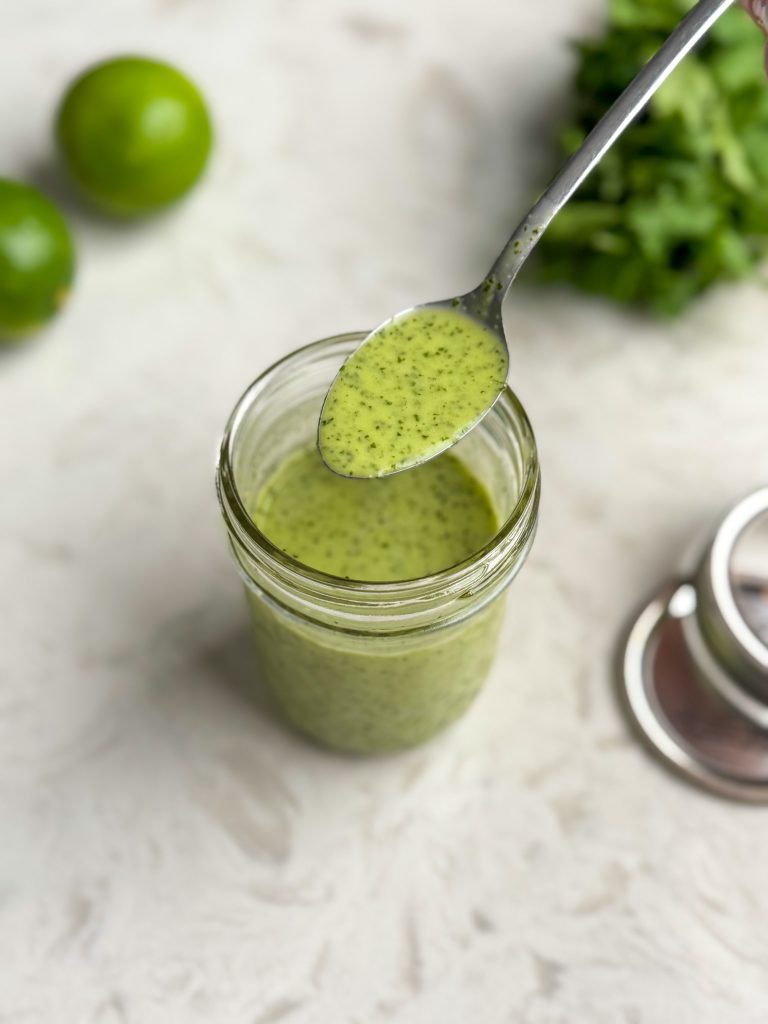 Spoonful of cilantro lime dressing in a glass mason jar with lid and cilantro and limes in the background.