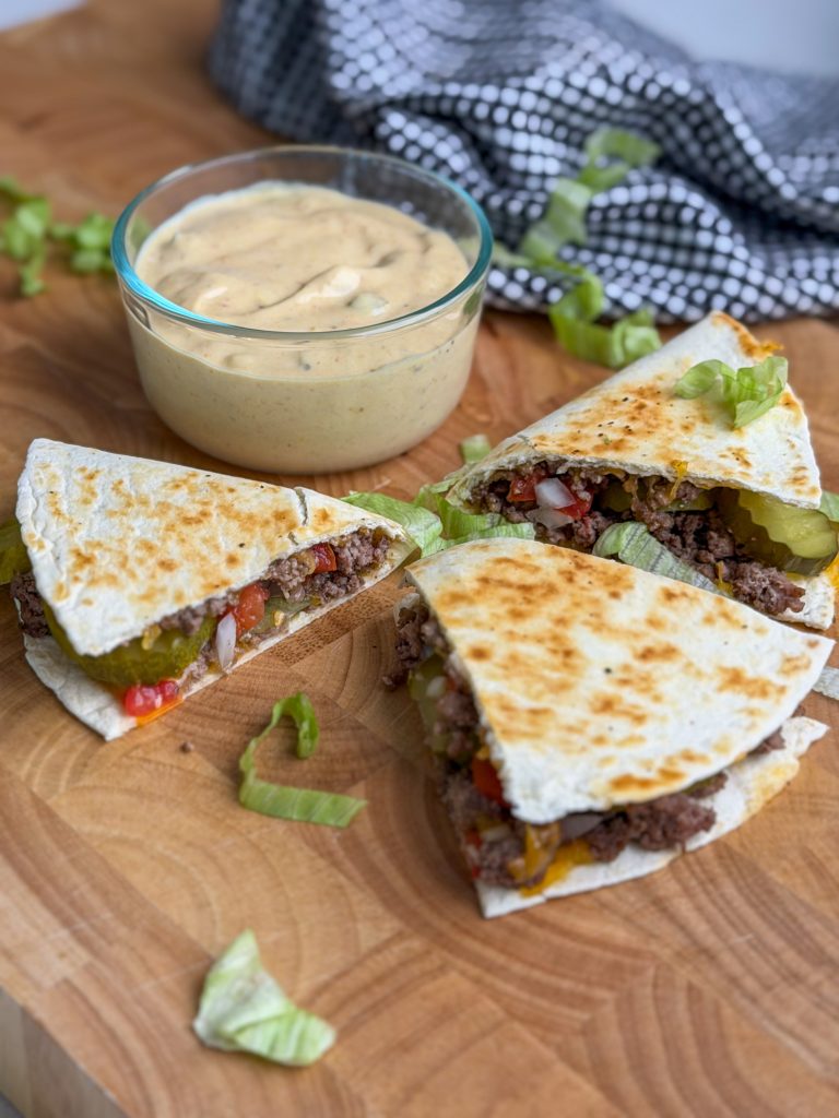 Big Mac quesadilla with tomato, pickles, lettuce, and light burger sauce on a brown cutting board.
