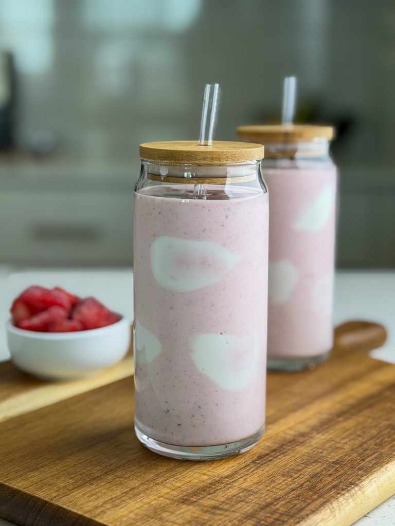 Strawberries and cream protein smoothie in a tall glass cup with straw and lid.