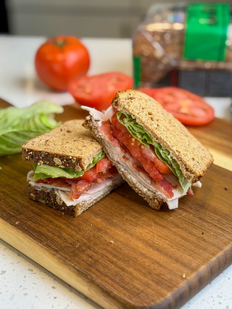 A turkey BLT sandwich cut in half on a dark brown wooden cutting board.