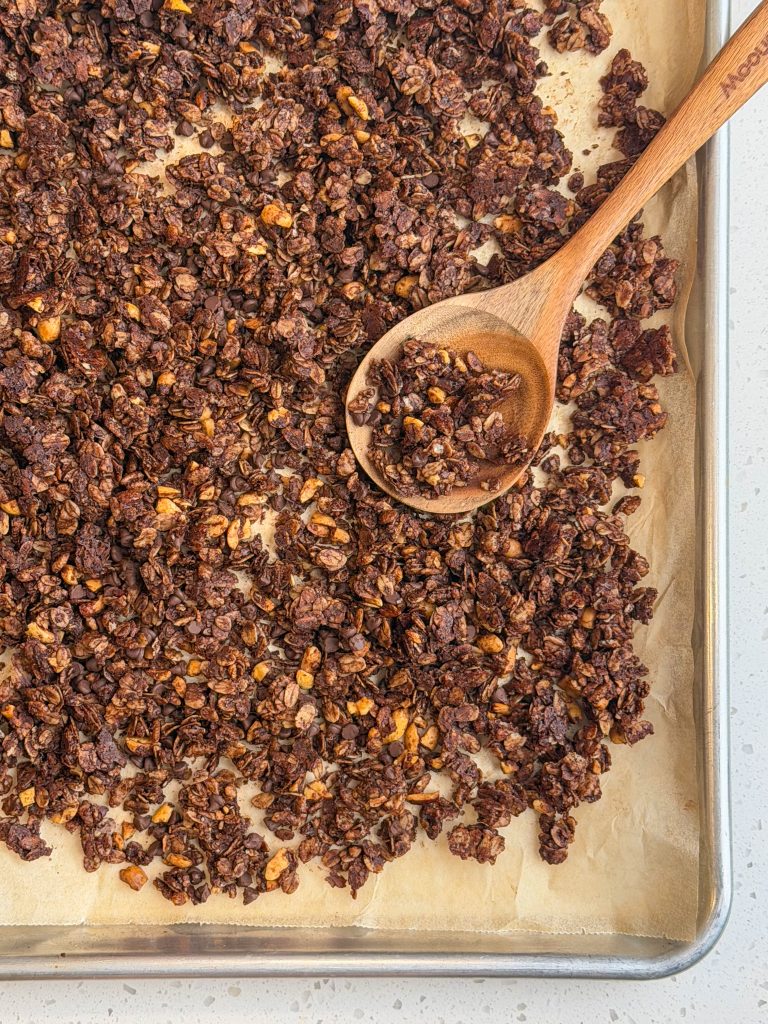 Chocolate crunch granola on a parchment lined sheet pan with a wooden spoon.